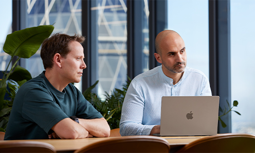 Ahmed, con su MacBook delante, sentado en una mesa con un compañero y mirando a alguien fuera de plano.