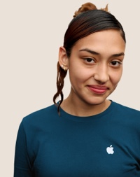 Portrait d’un membre de l’équipe Apple Retail souriant, avec de longs cheveux de couleur foncé et des mèches roses.