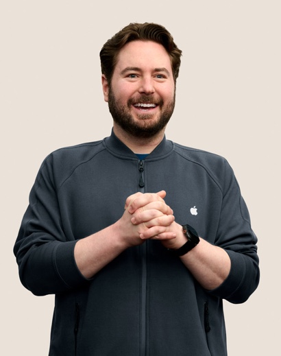 Portrait d’un membre de l’équipe Apple Retail souriant, avec des cheveux courts ondulés de couleur foncée et une barbe.