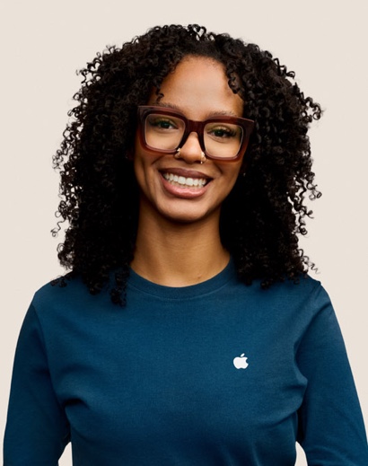 Portrait d’un membre de l’équipe Apple Retail souriant, avec de longs cheveux bouclés de couleur foncée et des lunettes.