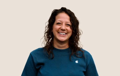 Portrait d’un membre de l’équipe Apple Retail souriant, avec de longs cheveux ondulés de couleur foncée.