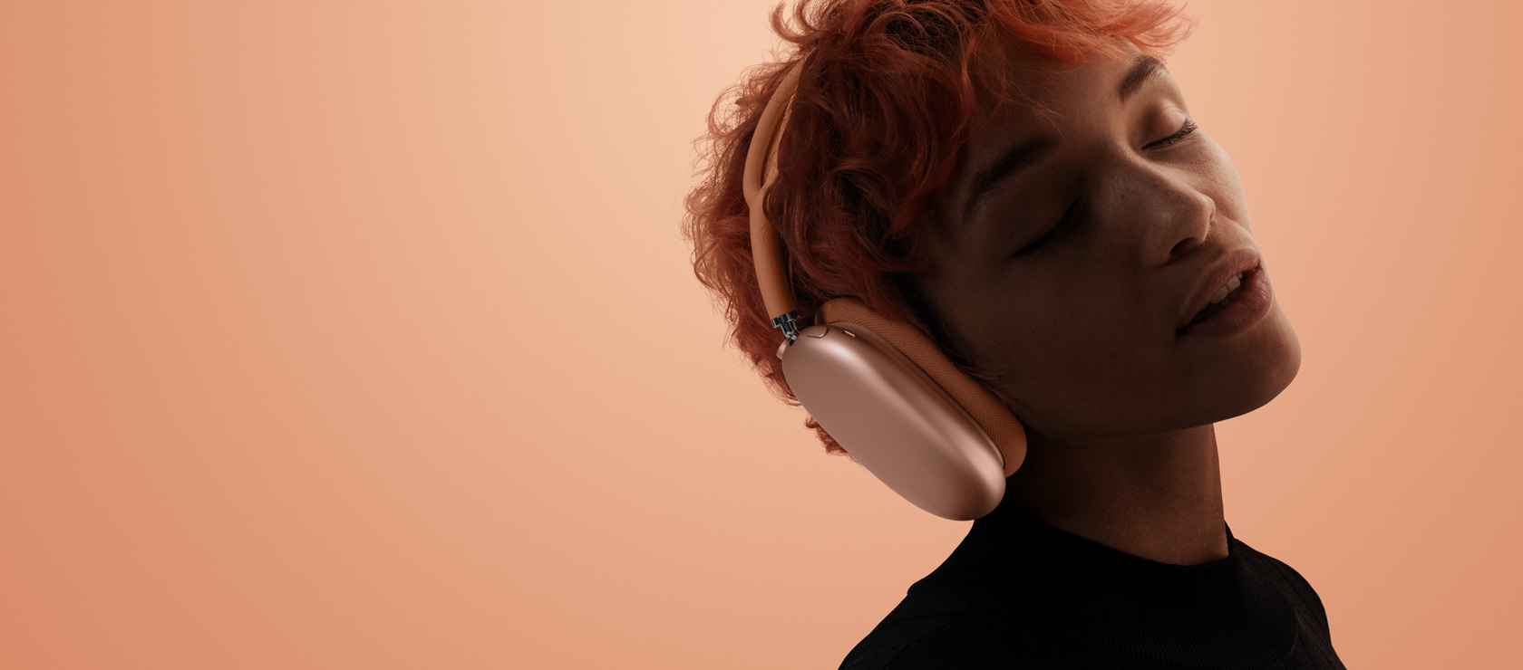 شخص رأسه مائل وعيناه مغلقتان يضع AirPods&nbsp;Max&nbsp;2 على رأسه، لون برتقالي، على خلفية بلون برتقالي وردي