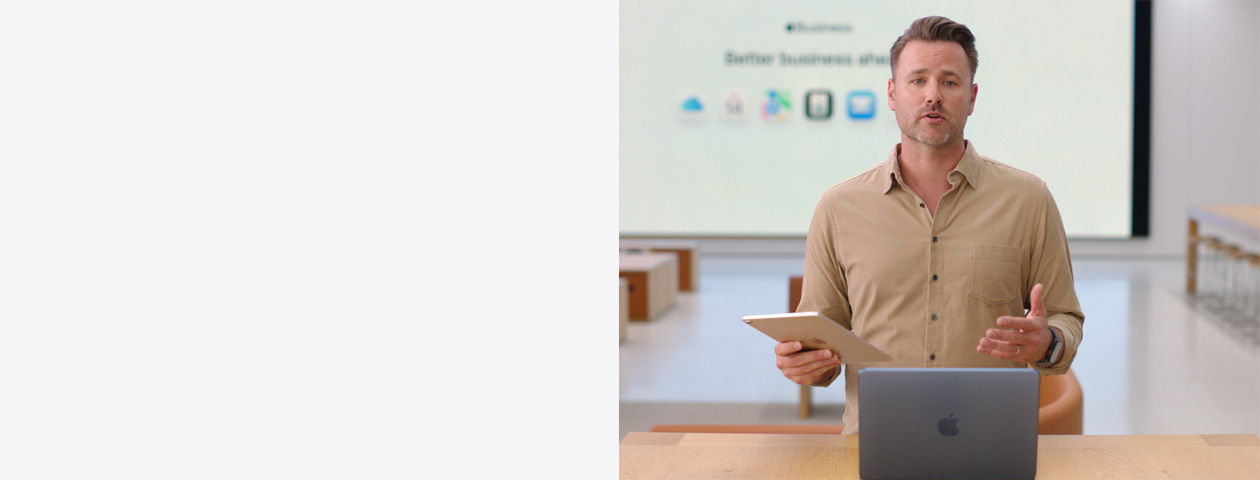 Présentateur parlant avec un iPad à la main, debout devant un MacBook posé sur une table, tandis qu’un grand écran derrière lui affiche les icônes de plusieurs apps professionnelles.