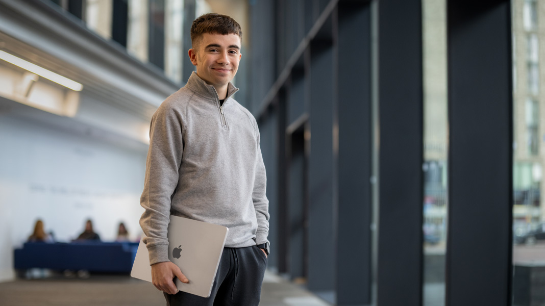 En leende student står i en korridor i en modern byggnad med en bärbar MacBook under armen