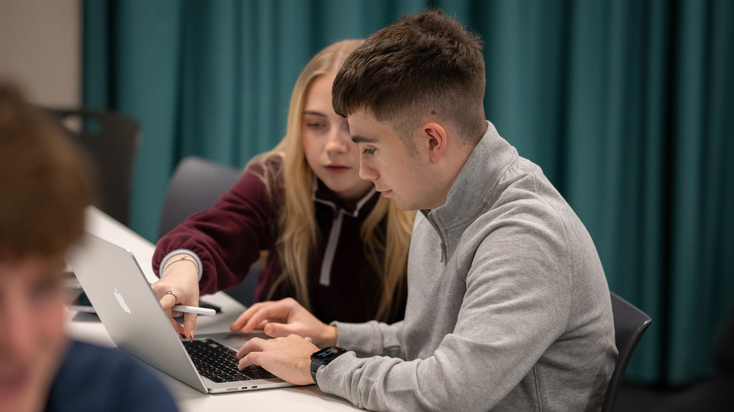 Två klasskamrater samarbetar på en MacBook, den ena pekar på skärmen medan den andra skriver