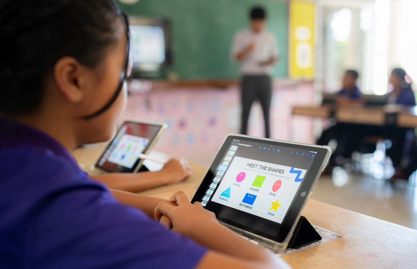 Gros plan d’une élève à son bureau, utilisant un iPad. À l’écran, des figures colorées s’affichent avec le titre « Meet the shapes ».