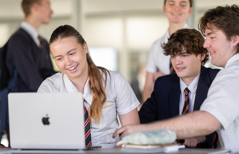 En grupp glada elever i skoluniform samlas runt en MacBook för att samarbeta.