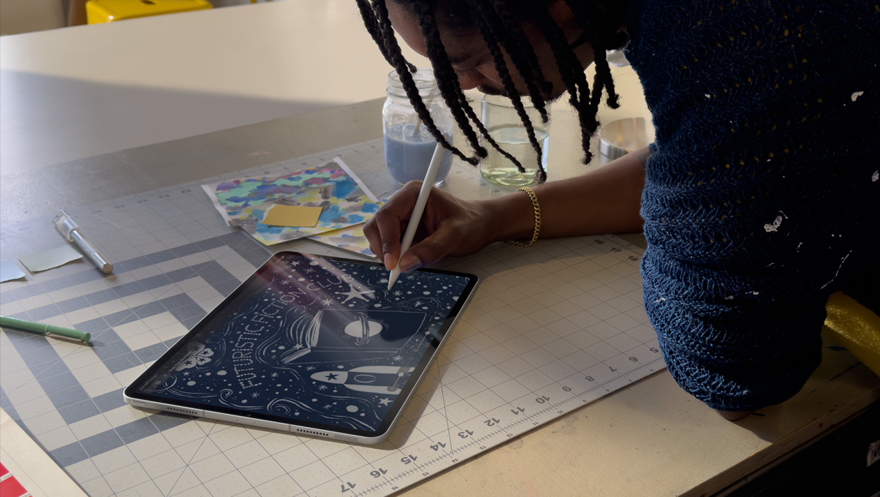 Personne avec des tresses portant un pull bleu qui dessine sur un iPad Pro avec un Apple Pencil, l’écran montre l’illustration d’un ciel nocturne avec une planète sur la couverture d’un livre, un vaisseau spatial, un papillon et futuristic fiction club écrit au centre